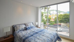 Une chambre avec un grand lit, une table de chevet, une fenêtre donnant sur des immeubles et une rue.