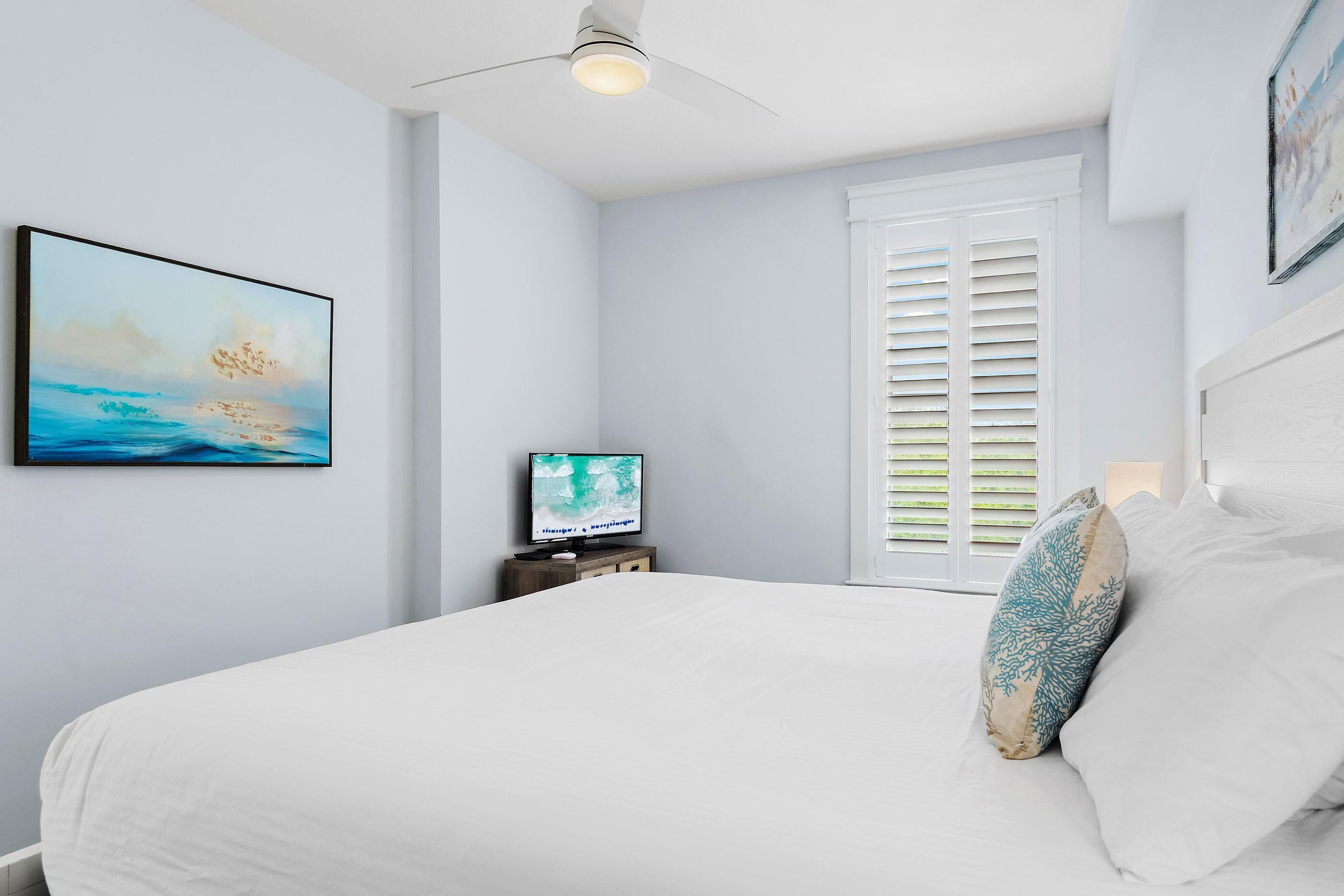 A bedroom with a bed, a television on a stand, a ceiling fan, and a window with shutters.