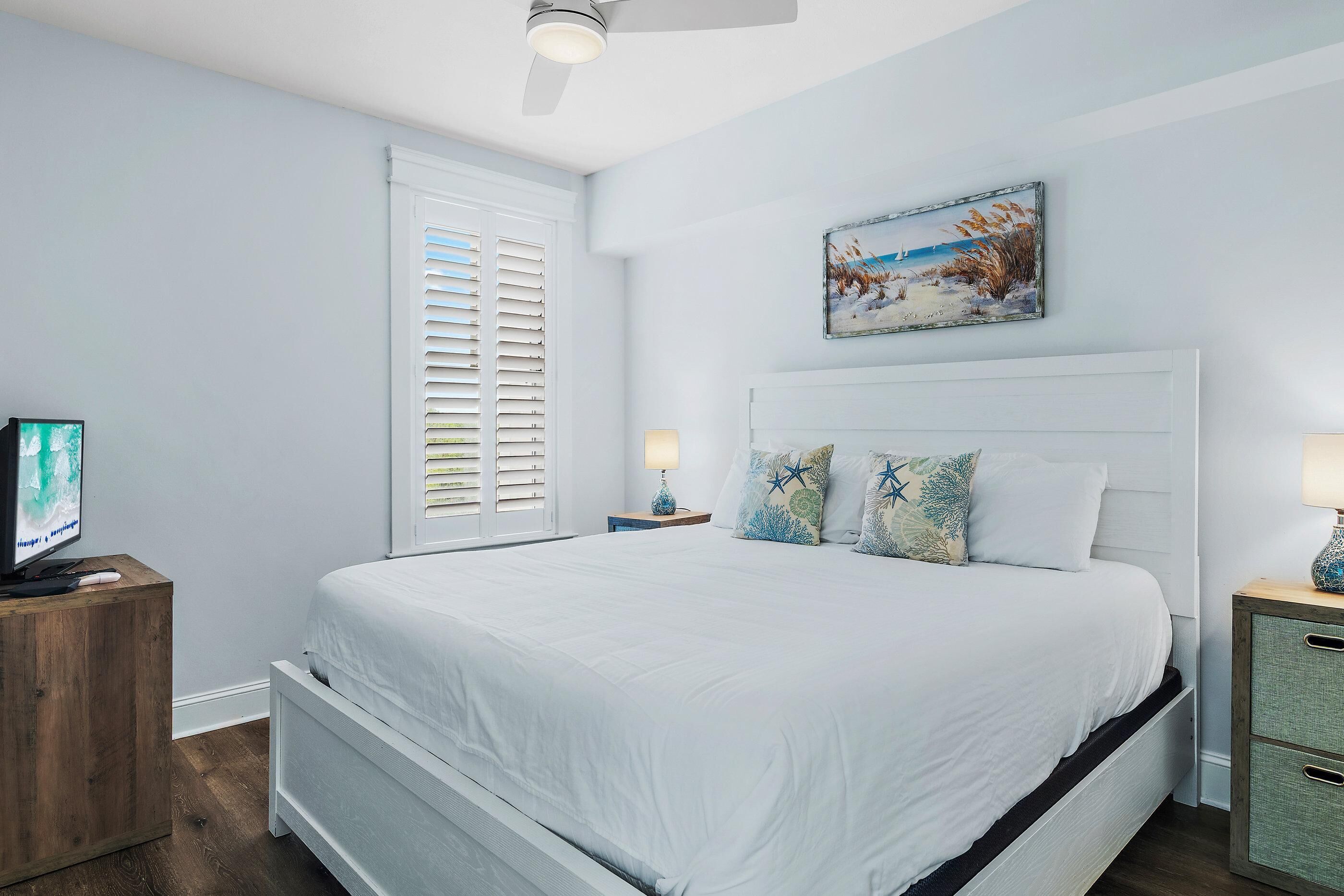 A bedroom with a bed, a nightstand, a TV, a painting, and a window with blinds.