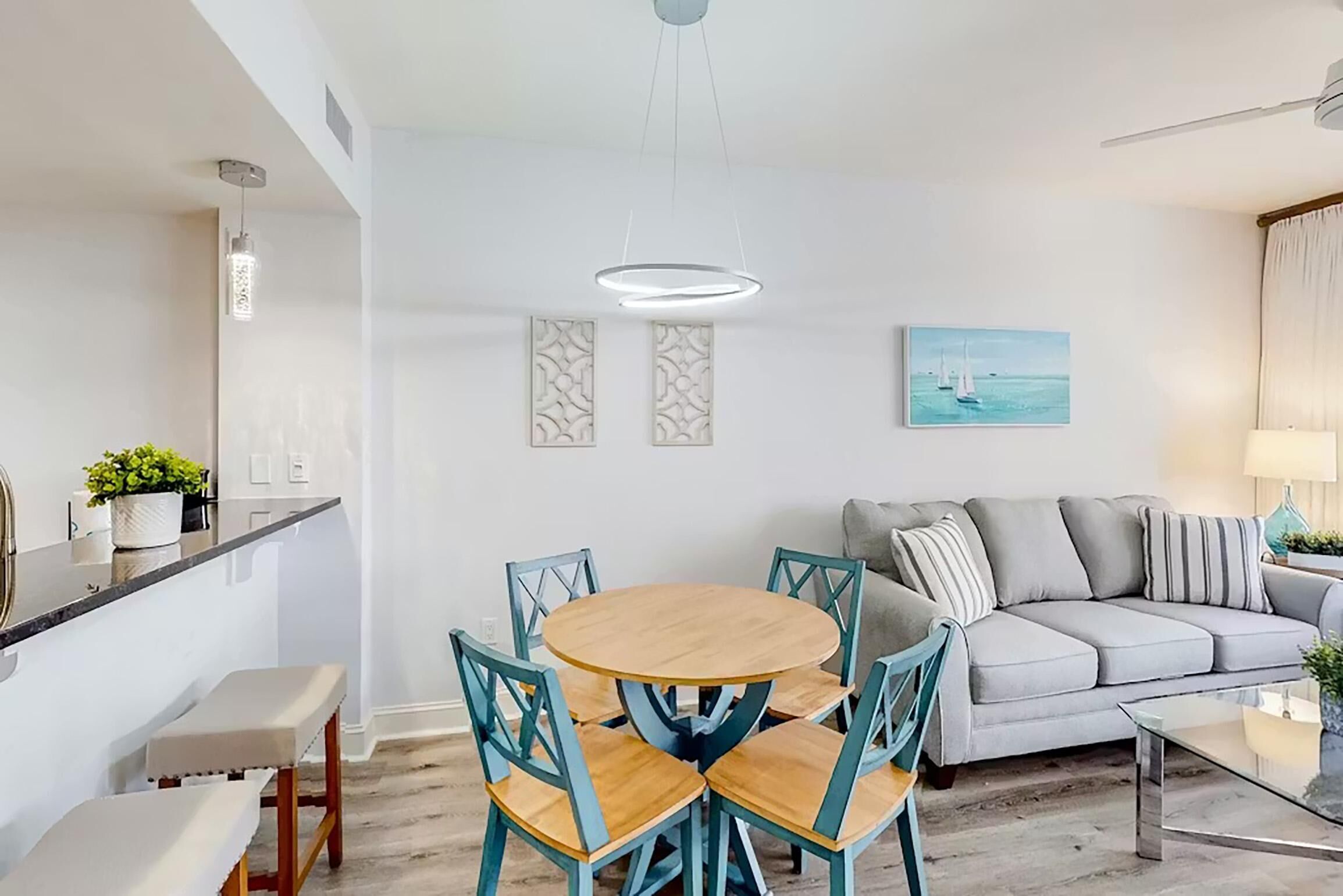 A modern living room with a round wooden table, teal chairs, a grey sofa, and a ceiling fan.
