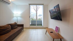 A modern living room with a sofa, a table, a TV mounted on the wall, and a large window with a view of buildings. - Zona Médica - Christus Muguerza - 2 Rec / 2 baños (Monterrey)