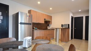 A modern kitchen with wooden cabinets, a stainless steel refrigerator, and a microwave. There are two round tables with grey mats, and a black door is visible in the background. - Zona Médica - Christus Muguerza - 2 Rec / 2 baños (Monterrey)