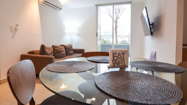 A modern living room with a glass-top dining table, brown sofa, and a TV mounted on the wall. - Zona Médica - Christus Muguerza - 2 Rec / 2 baños (Monterrey)