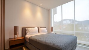 A bedroom with a large bed, a nightstand, a lamp, and a window with a city view. - Zona Médica - Christus Muguerza - 2 Rec / 2 baños (Monterrey)