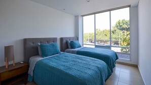 Quarto com duas camas, uma mesinha de cabeceira, abajur e uma janela grande com vista para árvores e um estacionamento.