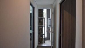 Un couloir étroit, avec une porte sur la droite, une fenêtre sur la gauche et une étagère métallique contenant divers objets.