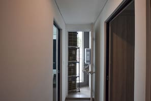 Um corredor estreito com uma porta à direita, uma janela à esquerda e uma estante de metal com objetos.