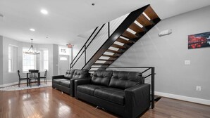 A modern living room with a black leather sectional sofa, a dining table with chairs, and a staircase with wooden beams.