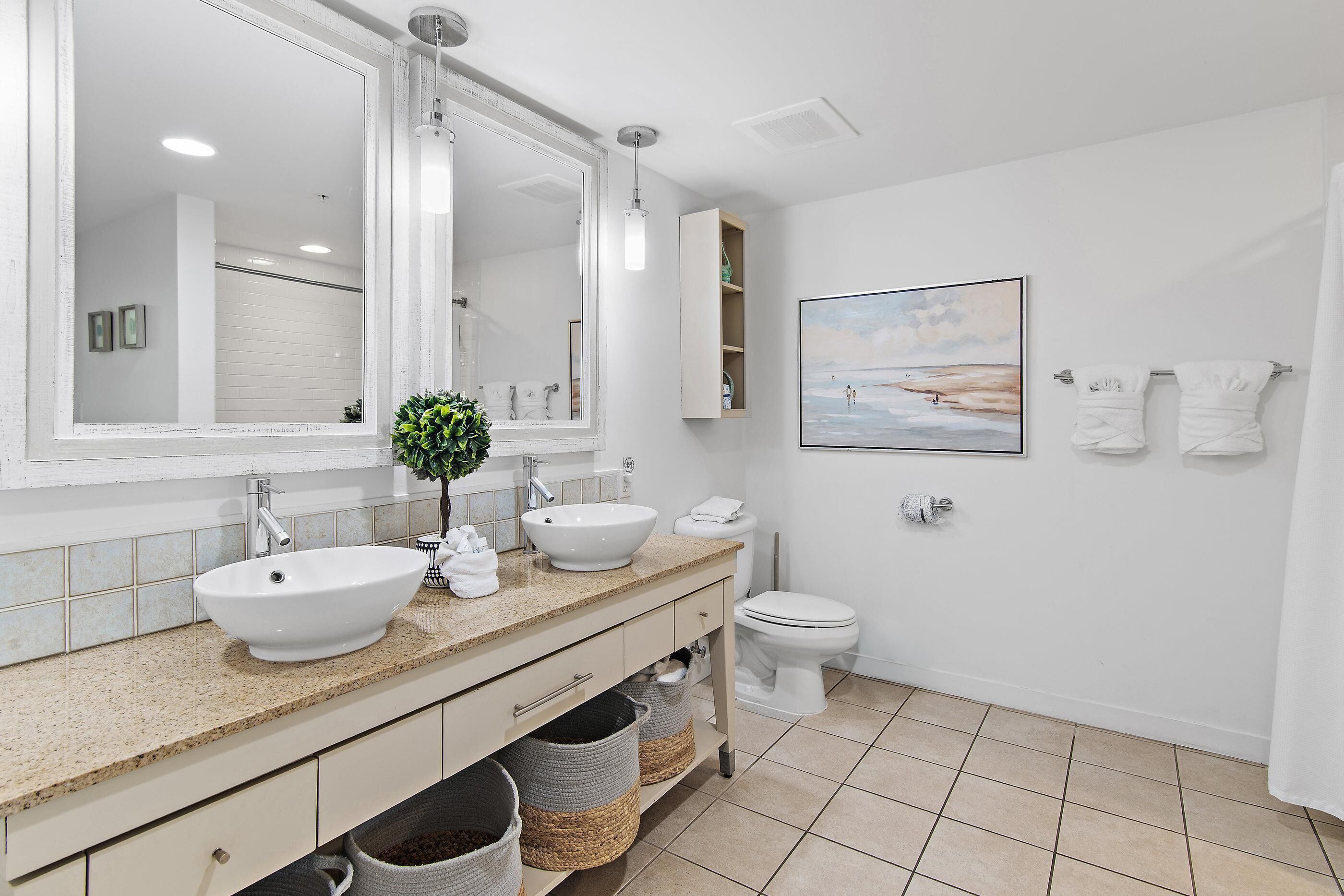 A bathroom with a large mirror, two sinks, a toilet, and a framed picture on the wall.