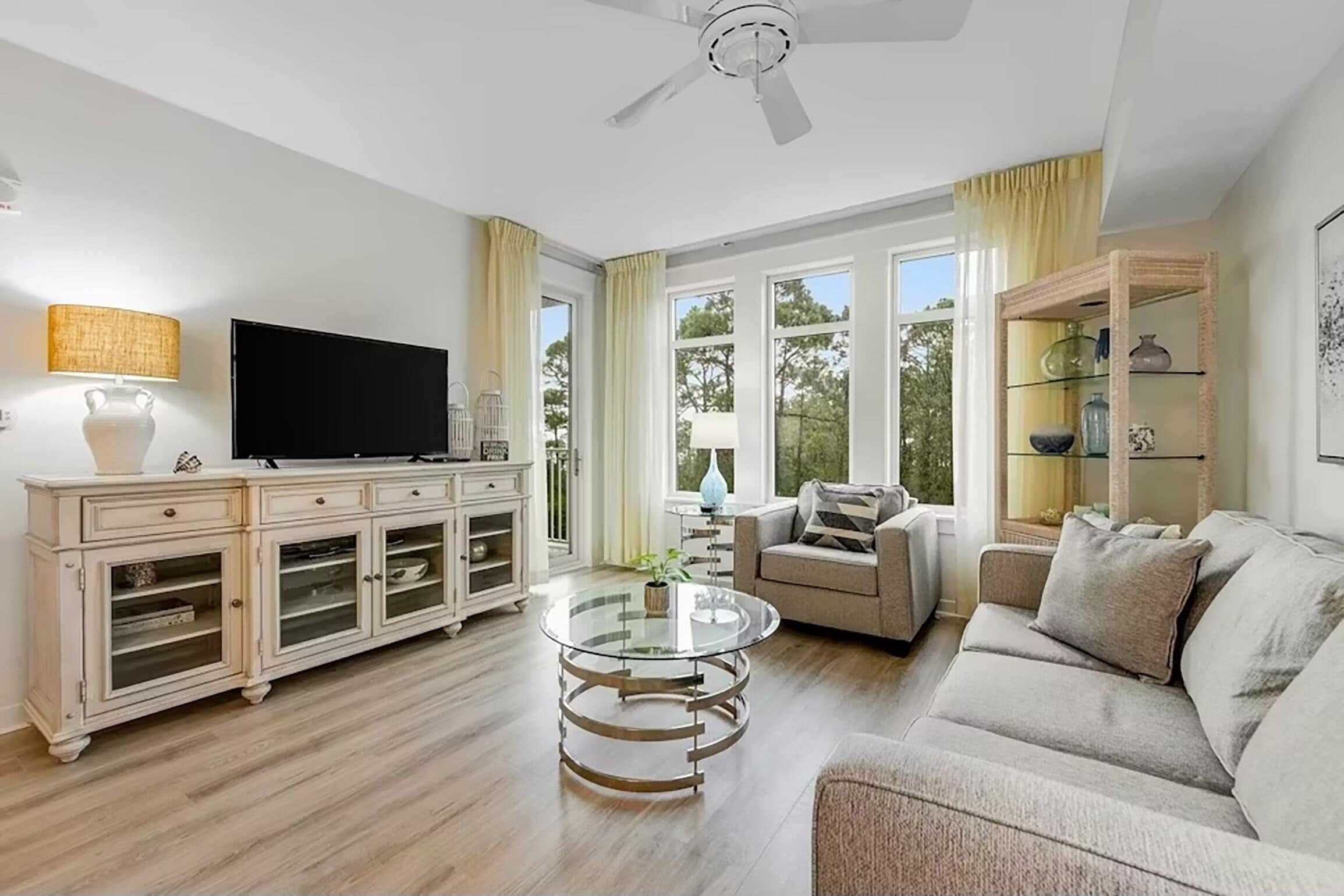 Una sala de estar con televisión, una mesa de centro de vidrio, un sofá, un mueble y un ventilador de techo.