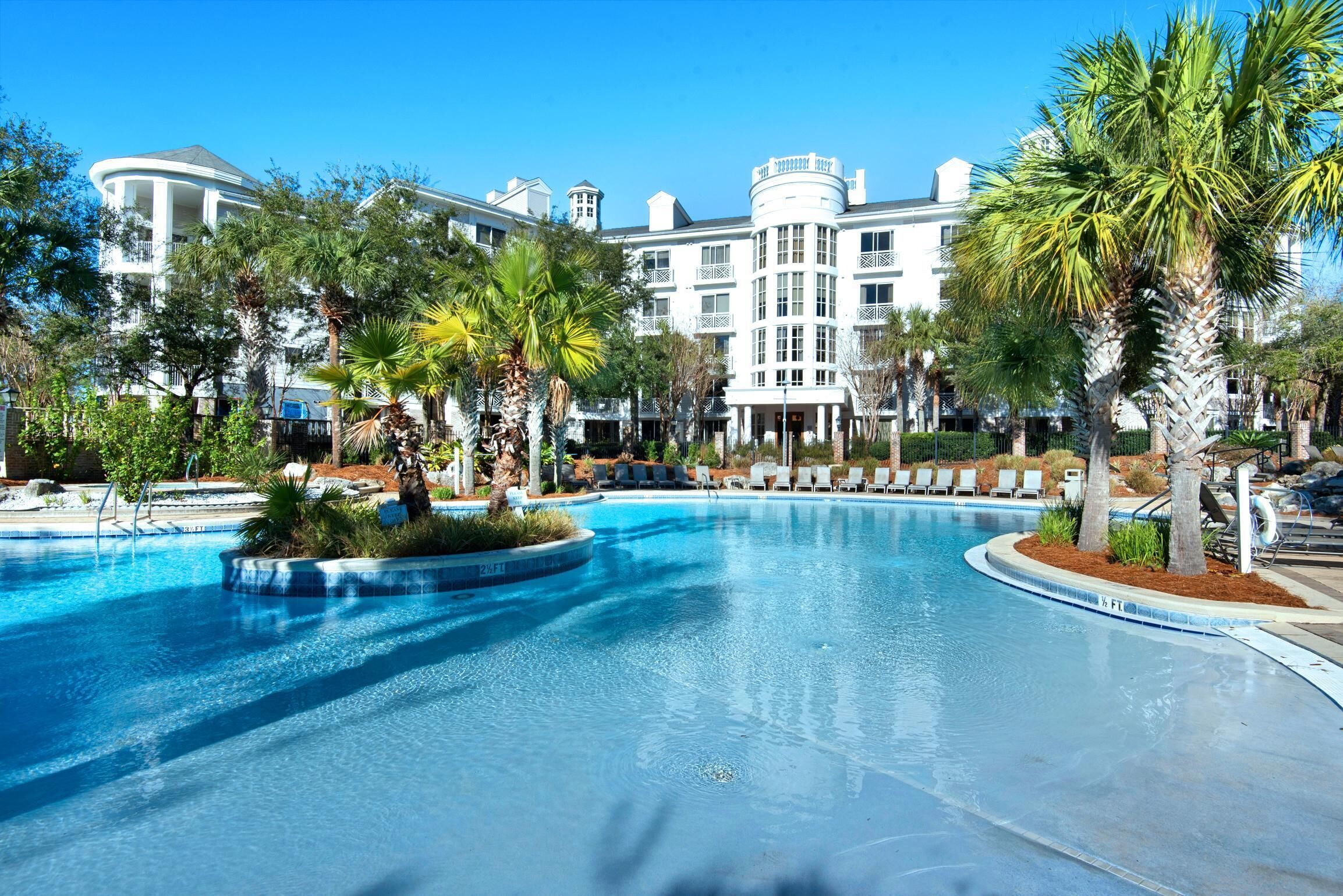 Una piscina amplia con sillas de descanso y palmeras, rodeada por un edificio tipo resort con varios balcones.