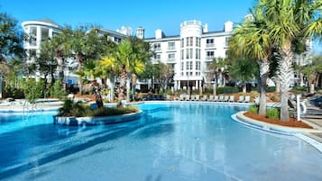 Una piscina amplia con sillas de descanso y palmeras, rodeada por un edificio tipo resort con varios balcones.
