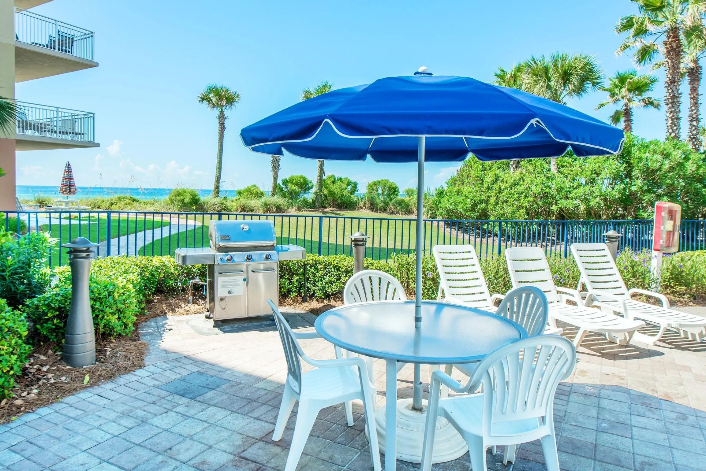 An outdoor patio with a grill, white chairs, and a blue umbrella.