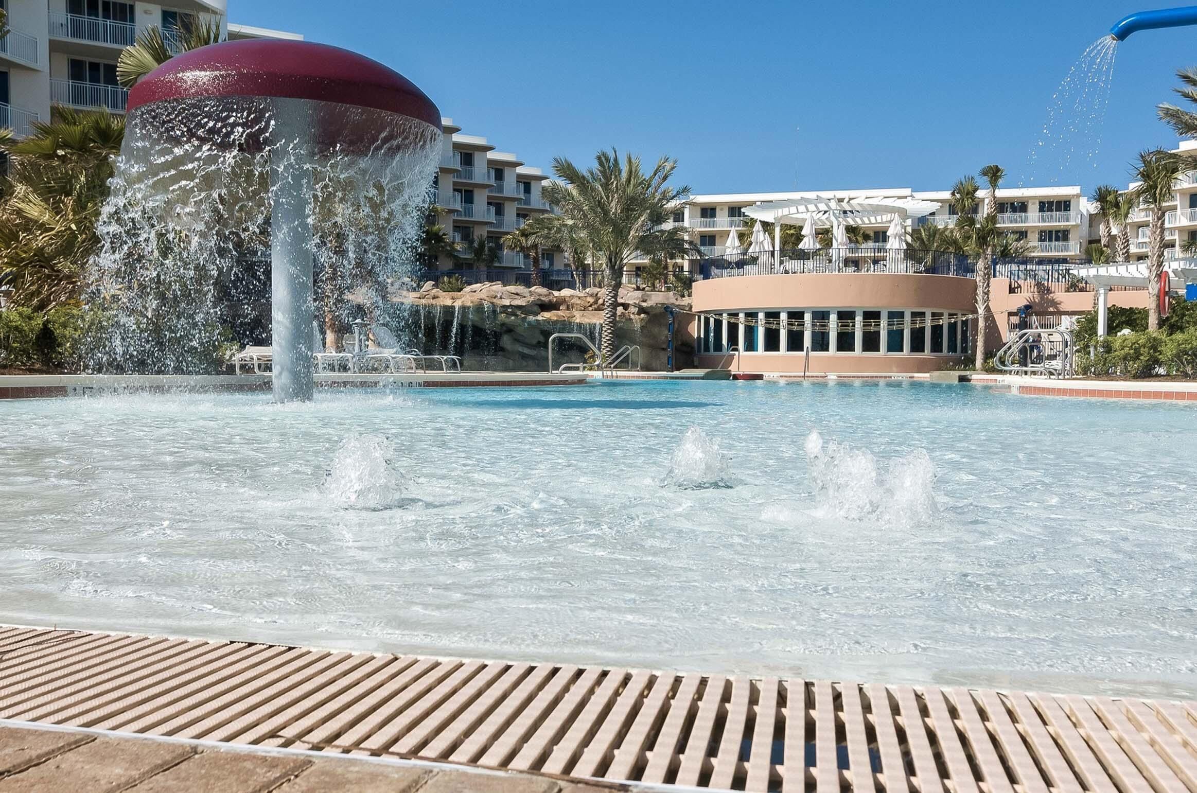 A pool with water jets, a palm tree, and a building with balconies.