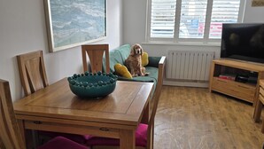 Ett matområde med ett träbord, stolar och en skål. Det finns en soffa med kuddar, en tv och ett fönster med persienner.