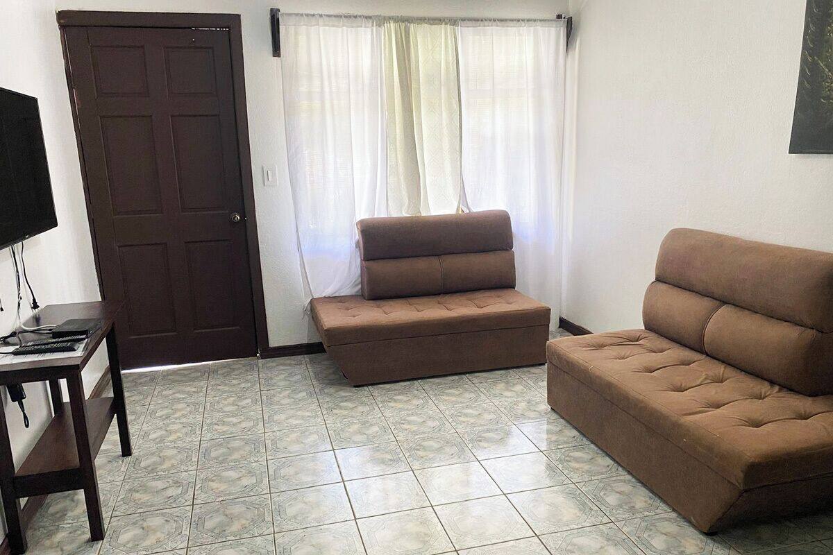 A room with a brown sofa, a small table with a keyboard, a closed door, and a window with sheer curtains.