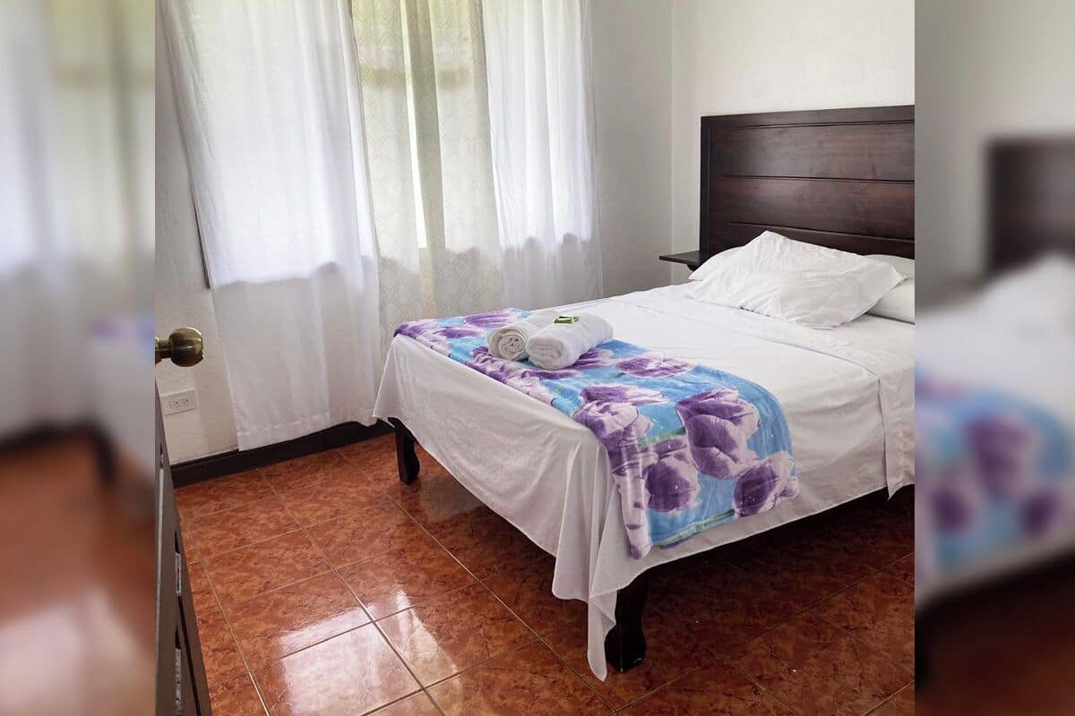 A hotel room with a bed, a nightstand, a window with curtains, and a tiled floor.