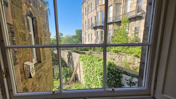 Ein Fenster mit Blick auf eine Steinmauer und Grünpflanzen, ein kleiner Tisch mit einem Blumenstrauß sowie ein Gebäude mit mehreren Fenstern und Balkonen.
