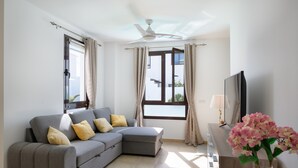 Ein modernes Wohnzimmer mit einem grauen Ecksofa, einem Flachbildfernseher, einem Deckenventilator und einem Fenster mit Vorhängen.