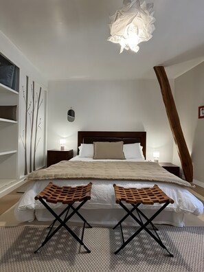 Un dormitorio con una cama, dos sillas de madera, una mesita de noche, una lámpara, un cuadro con árboles en la pared y una lámpara del techo.