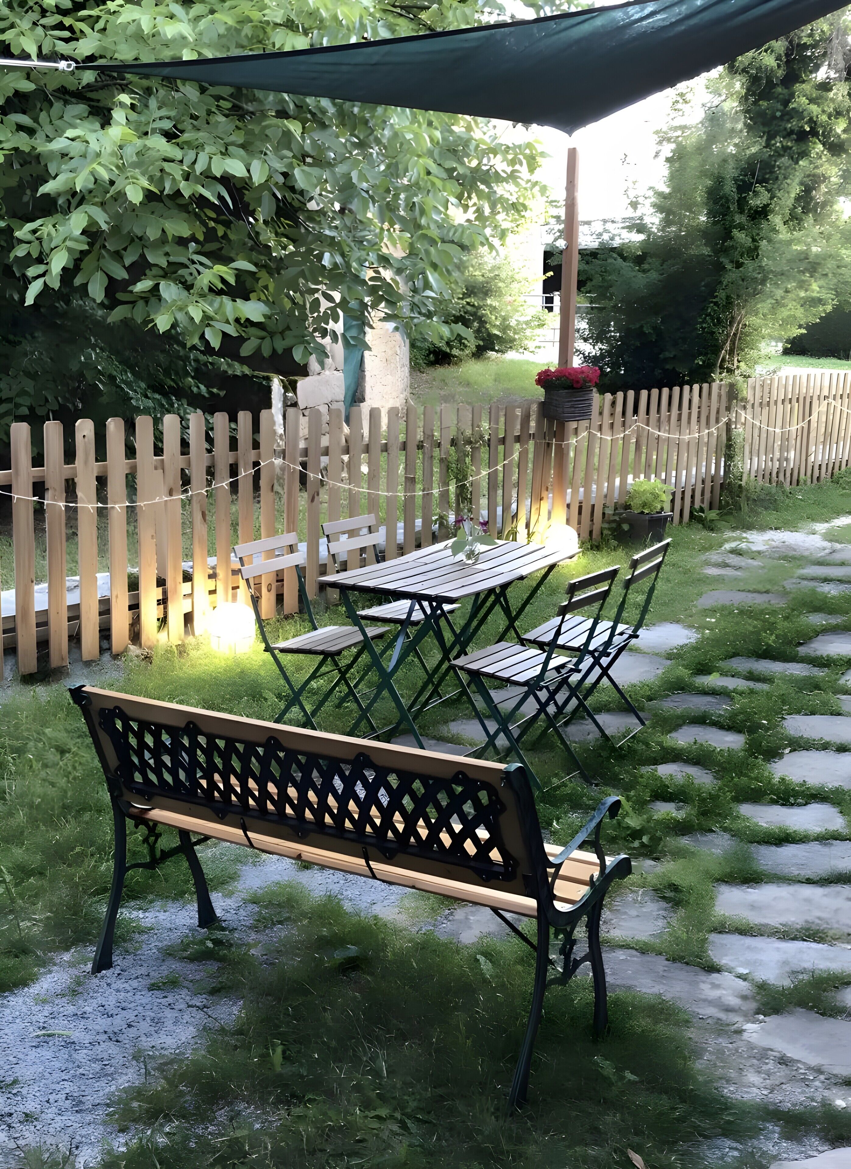 A garden patio with a wooden bench, metal table and chairs, a string of lights, and a wooden fence.