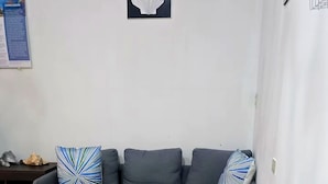 A living room with a grey sofa, teal armchair, and patterned cushions, a framed artwork on the wall, and a tiled floor.