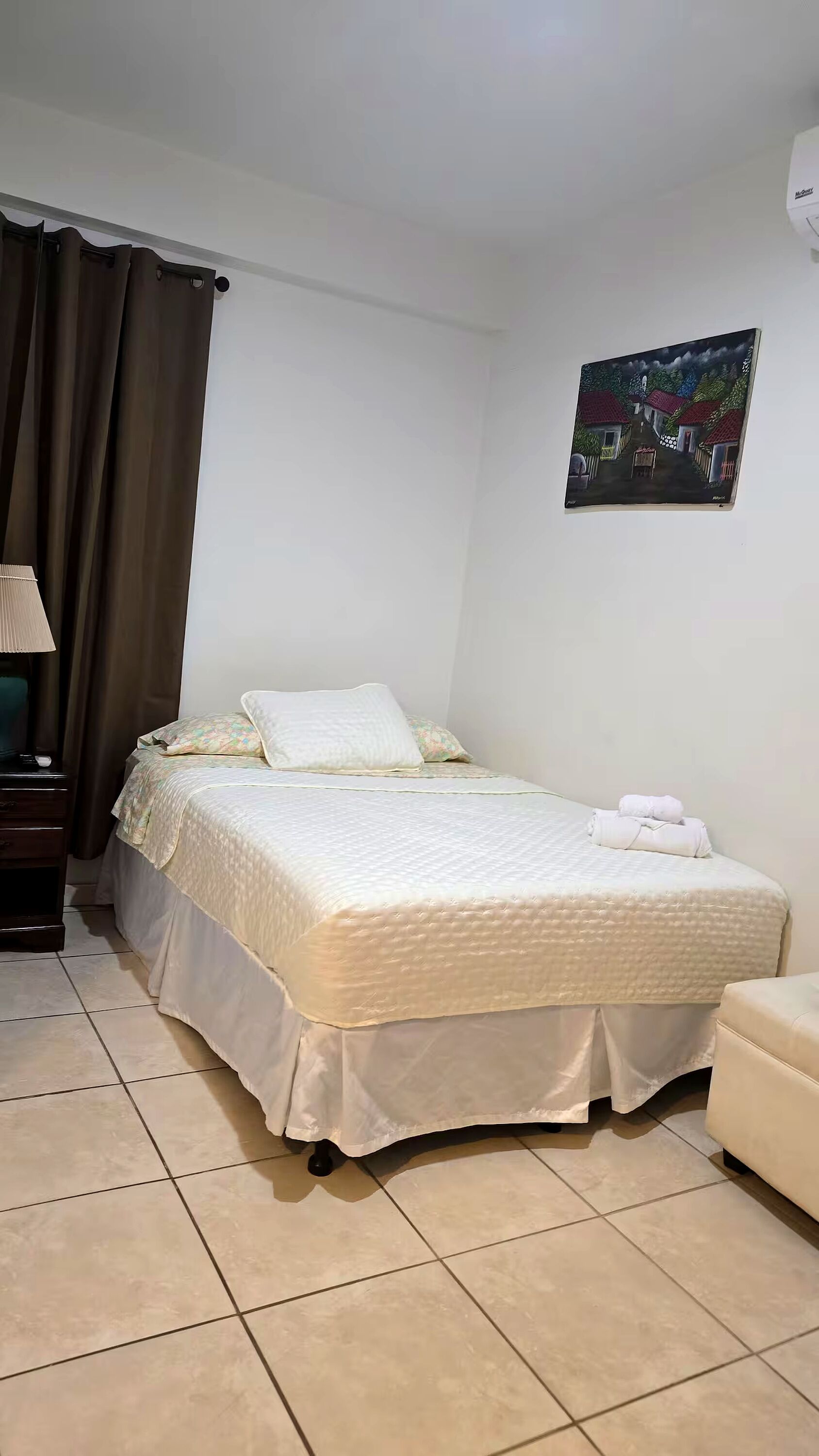 Un dormitorio ordenado con una cama, una mesita de noche, una lámpara, un cuadro en la pared y una silla.