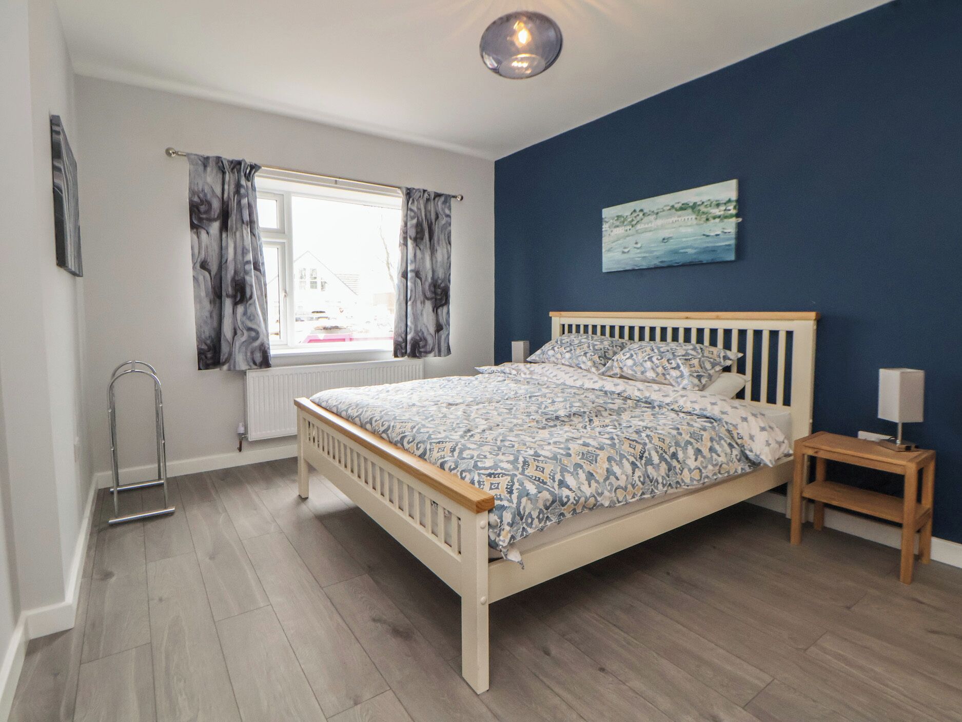 A bedroom with a wooden bed, a window with curtains, a radiator, a painting on the wall, and a bedside table with a lamp.