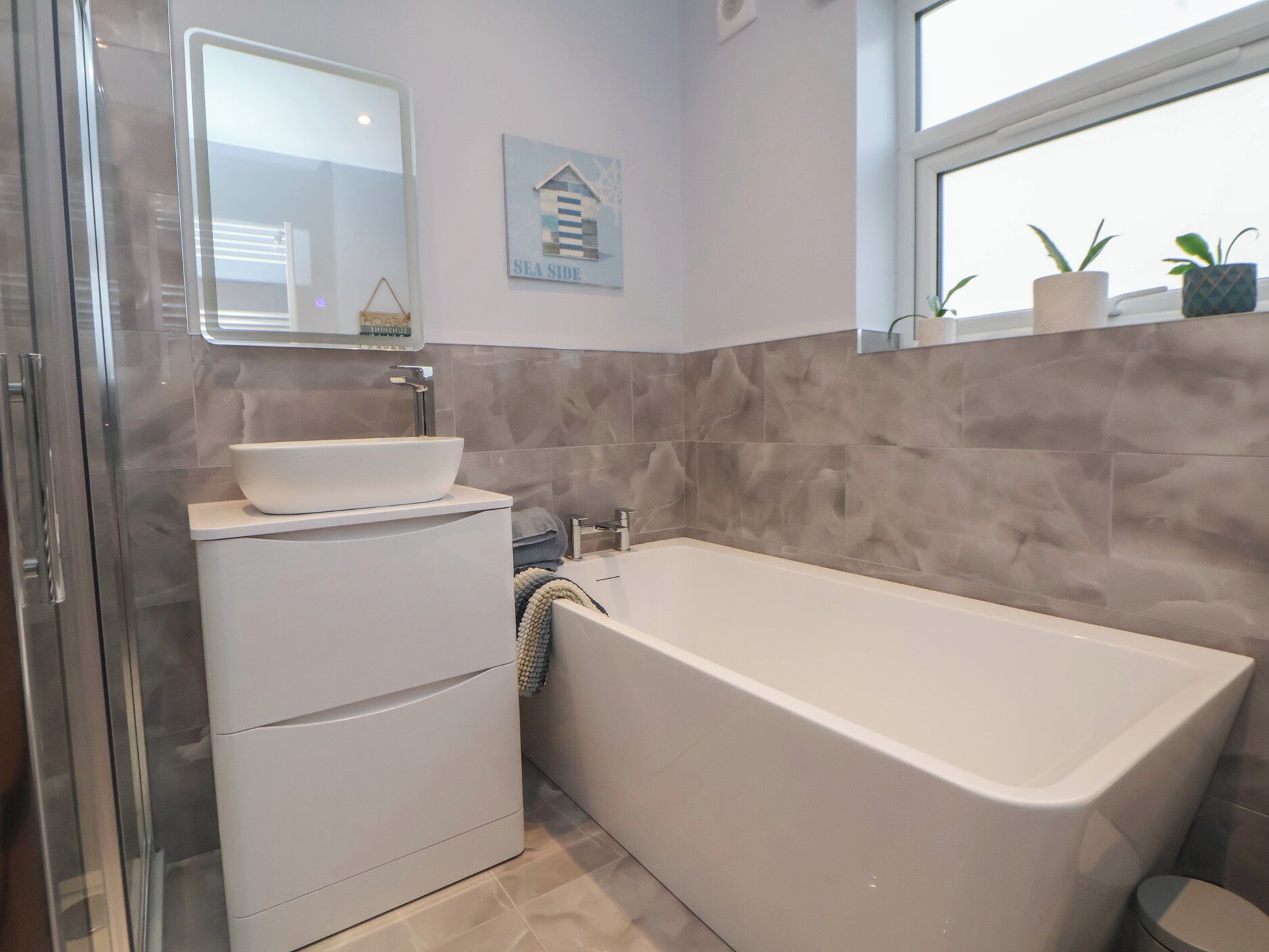 Ein Badezimmer mit einem weißen Waschbecken, einer Badewanne und einem Fenster mit Pflanzen.