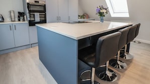 A modern kitchen with grey cabinets, a built-in oven, a central island with bar stools, and a window providing natural light. - Mikes Place On The Cove (Rhyl)