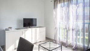 A modern room with a flat-screen TV on a white cabinet, a glass coffee table, and sheer curtains on a window. - T2 Vert Anis Apartment with Wi-Fi (Salies-de-Béarn)