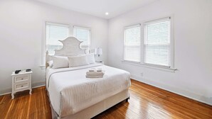 Quarto com cama, mesa de cabeceira e duas janelas com persianas.