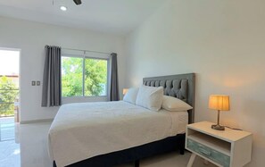 Quarto com cama, mesa de cabeceira com abajur, janela com cortinas e vista para o verde lá fora.
