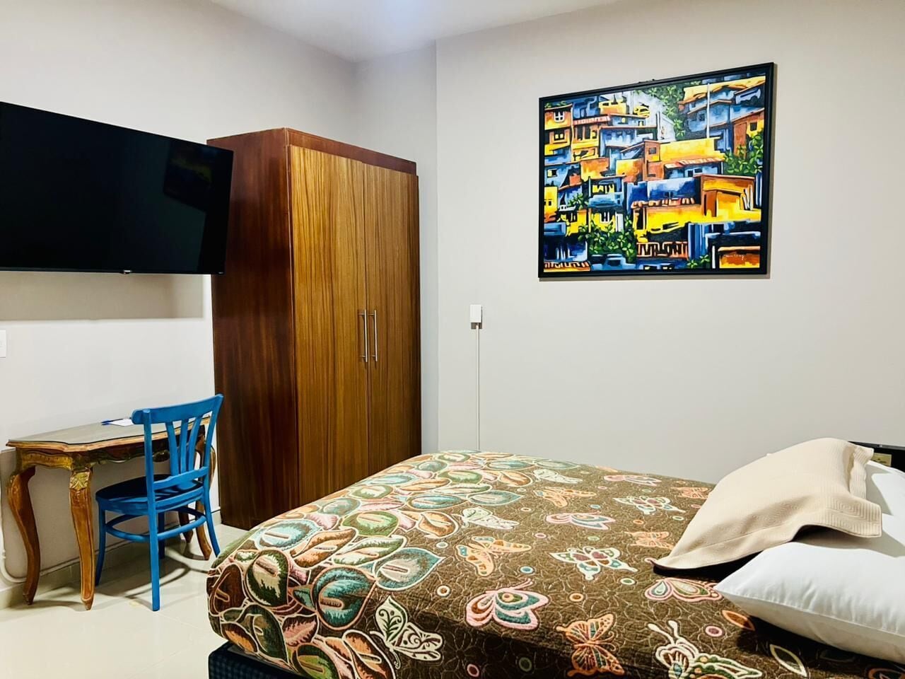 A bedroom with a bed, a desk, a chair, a wardrobe, and a painting on the wall.