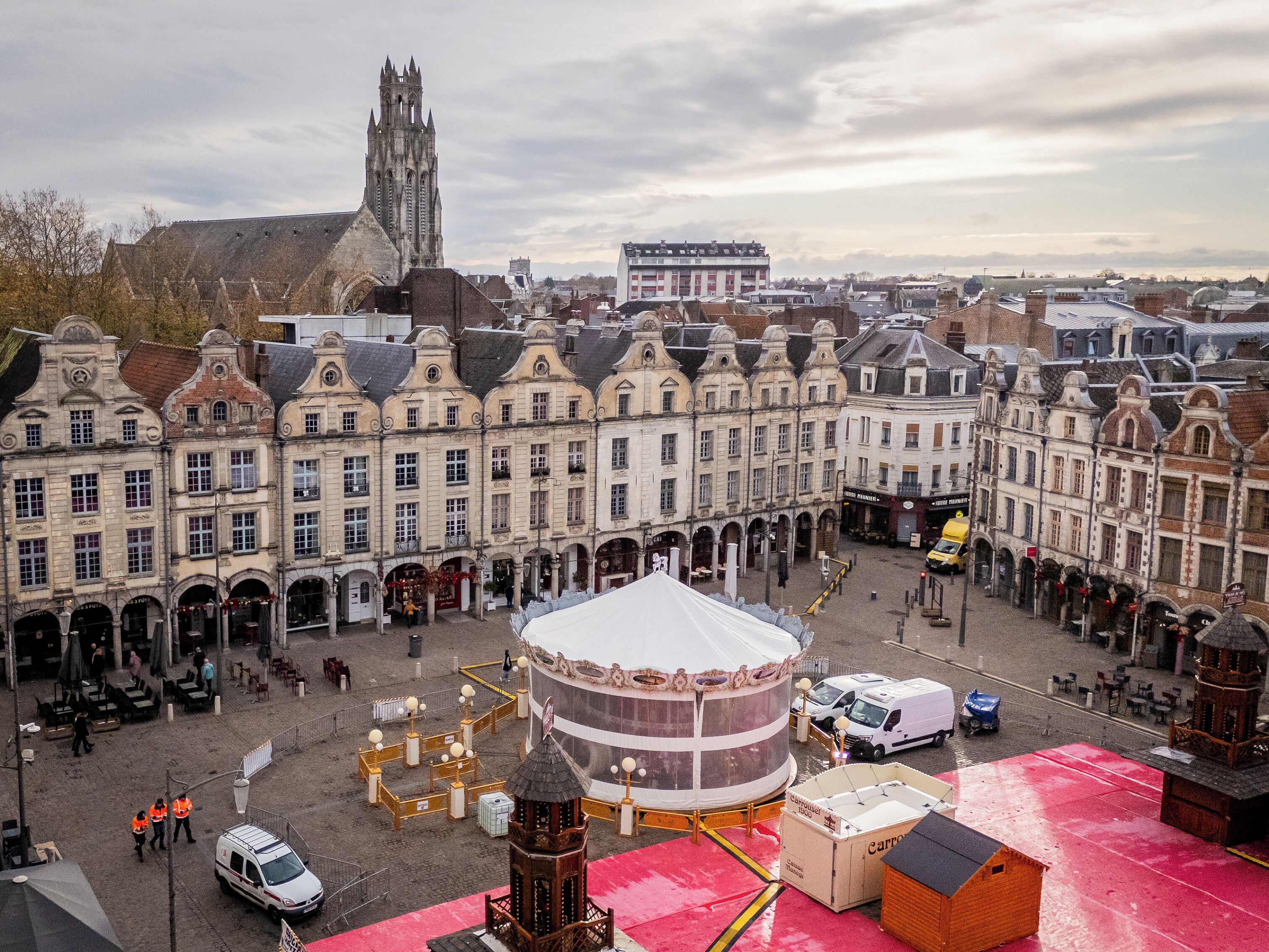Una piazza storica con architettura europea tradizionale, una torre campanaria imponente e uno spazio aperto centrale con una tenda bianca.