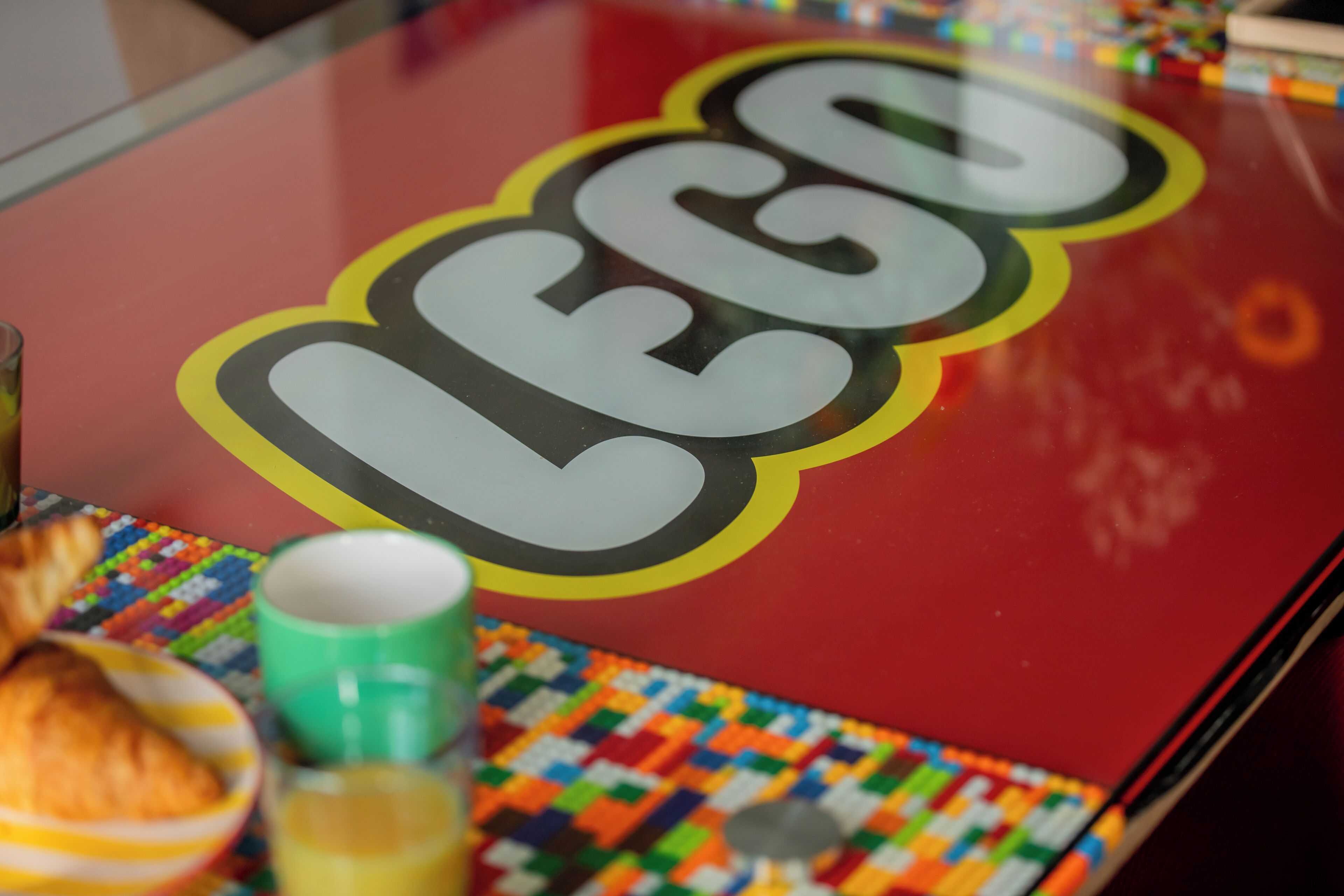 A table with a LEGO logo, a green mug, a plate with a croissant, and a colorful tablecloth.