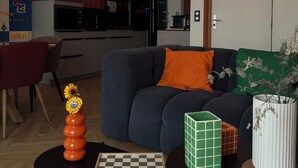 A modern living room with a sofa, a chessboard, a vase with flowers, and a kitchen in the background.