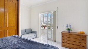 A bedroom with a bed, a wooden dresser, a grey sofa, and a sliding glass door leading to a balcony. - Holiday Apartment 'Panoramic View Apartment' with Sea View, Wi-Fi and Air Conditioning (Quarteira)