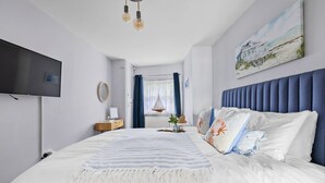 A bedroom with a large bed, a flat-screen TV, a wooden nightstand, a sailboat decoration, and a painting of a beach house. - Beach View Suite - Sea view, Balcony, Wi-fi (Torquay)