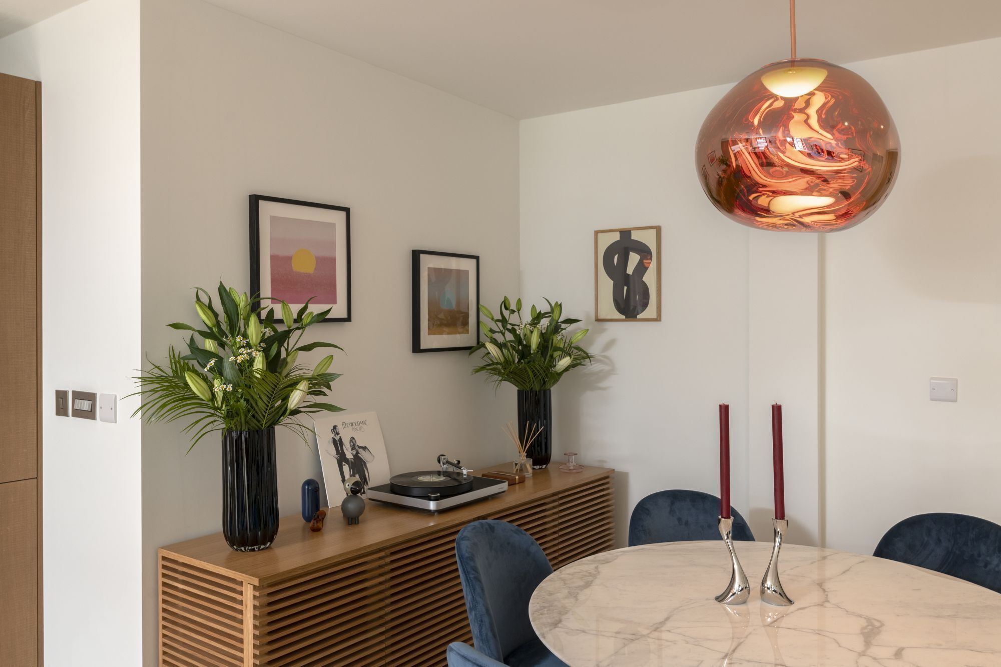 A modern dining area with a round marble table, blue chairs, a wooden sideboard, and two potted plants.