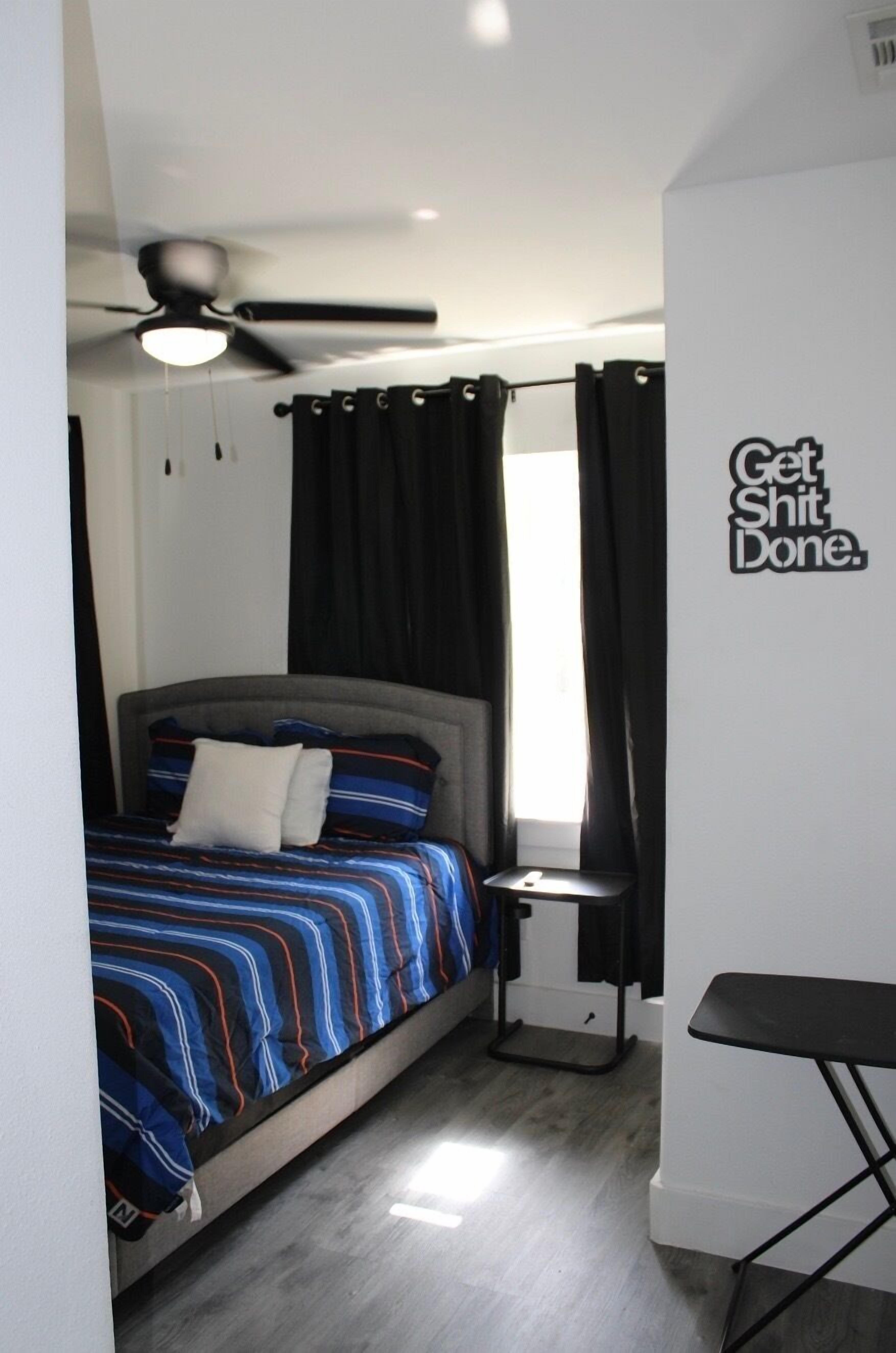 A bedroom with a bed, a ceiling fan, a window with curtains, a small table, and a motivational wall quote.