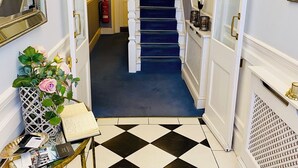 Un pasillo con suelo a cuadros blanco y negro, una escalera con alfombra azul, una puerta blanca y un espejo a la izquierda.