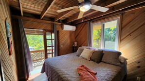 A wooden cabin bedroom with a bed, ceiling fan, and large windows. - Sandy Bay Island Retreat w/Private Dock, Reef Snorkeling, Pool & Walk to Beach (Roatán)