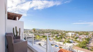 Ein Balkon mit Korbmöbeln, einem Kaktus und Blick auf eine Stadtlandschaft mit Gebäuden und Grünflächen.