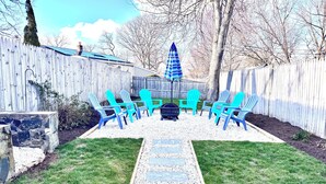 A backyard with a stone pathway, a fire pit, and blue Adirondack chairs arranged around a table.