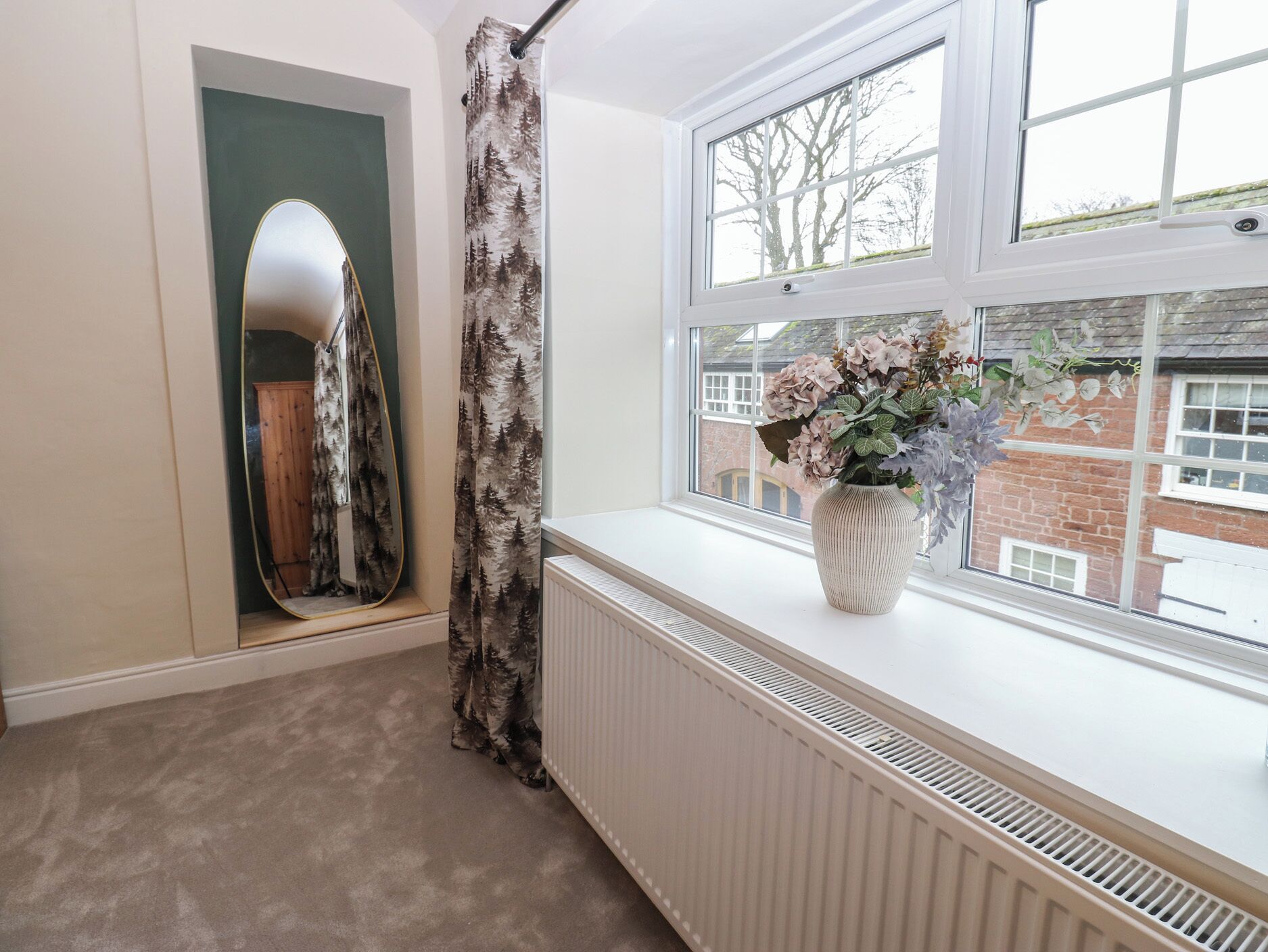Une pièce avec une grande fenêtre, un radiateur, un miroir et un vase de fleurs posé sur le rebord de la fenêtre.