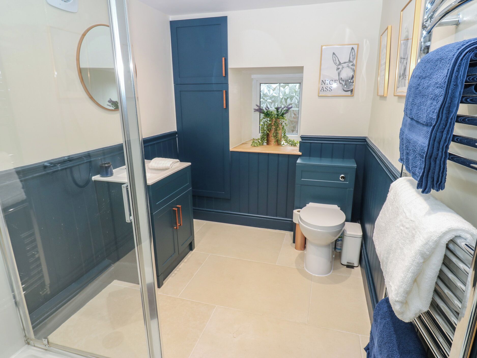A bathroom with a toilet, sink, and mirror, featuring blue cabinetry and a window with a plant.