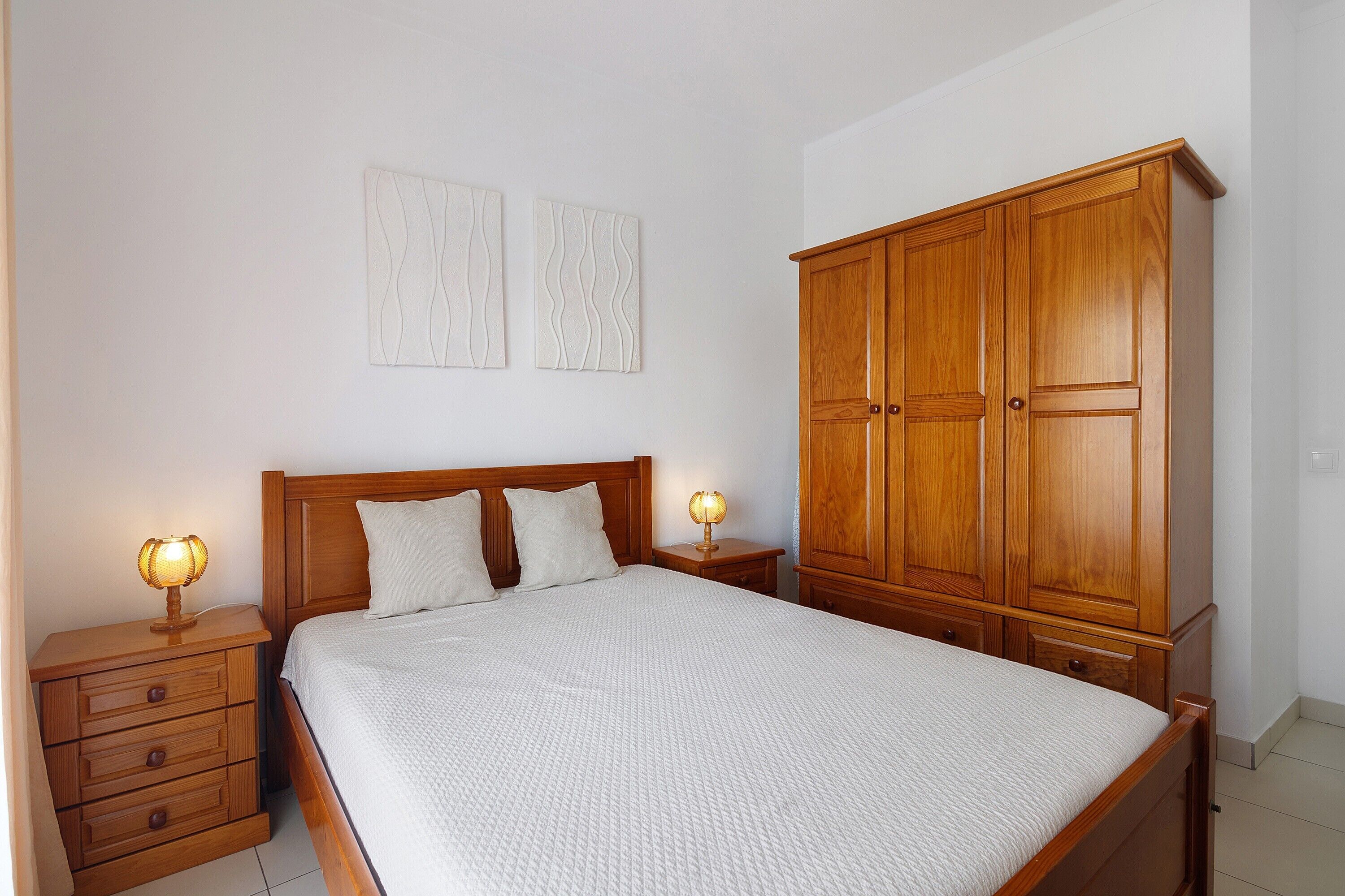 A bedroom with a wooden bed, two bedside tables with lamps, a wardrobe, and two wall-mounted artwork pieces.