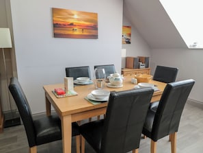A dining area with a wooden table, black leather chairs, a painting of a sunset, and a small kitchen area in the background. - Sunset Point (Alnwick)