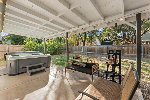 A covered patio with a hot tub, outdoor furniture, and a wooden fence in the background. - Spacious Home + Hot tub + Fire Pit + Play Loft (Cedar Park)